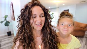 Mother with long curly hair and daughter with a bun smiling in a bright living room, indoors in a home setting.