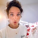 Close-up of a woman with wide eyes and surprised expression in a bright, white room, with a blood-stained blanket on the right.