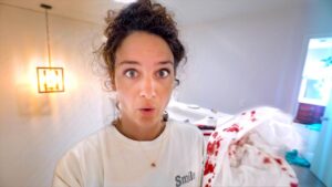 Close-up of a woman with wide eyes and surprised expression in a bright, white room, with a blood-stained blanket on the right.