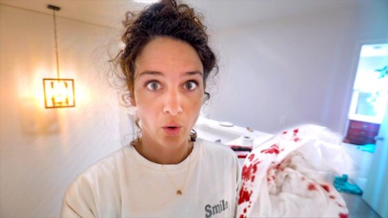 Close-up of a woman with wide eyes and surprised expression in a bright, white room, with a blood-stained blanket on the right.