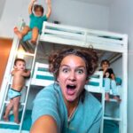 A woman takes a selfie in a kids' bedroom with a white bunk bed while several children play around, her mouth open in surprise/fun expression.