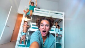 A woman takes a selfie in a kids' bedroom with a white bunk bed while several children play around, her mouth open in surprise/fun expression.