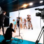 Behind-the-scenes of a studio photoshoot: kids posing in front of a white backdrop as crew adjust lighting and a large camera records nearby.