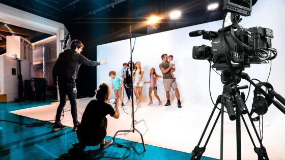 Behind-the-scenes of a studio photoshoot: kids posing in front of a white backdrop as crew adjust lighting and a large camera records nearby.