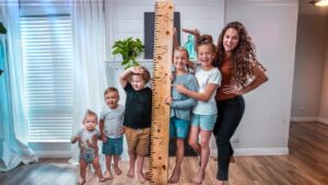 A woman and six children pose indoors with a wooden height ruler, measuring their heights together for a fun growth chart.