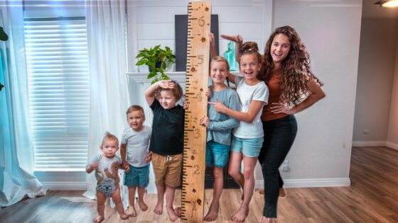 A woman and six children pose indoors with a wooden height ruler, measuring their heights together for a fun growth chart.