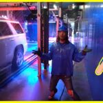 Person in blue rain gear and hard hat standing in a wet industrial hallway with a white SUV on a track nearby; colorful lights around.