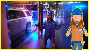 Person in blue rain gear and hard hat standing in a wet industrial hallway with a white SUV on a track nearby; colorful lights around.