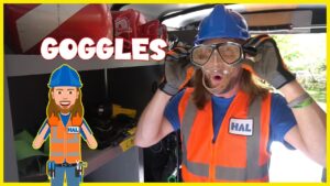 Person in a blue hard hat and orange safety vest wears clear protective goggles inside a workshop; the word 'GOGGLES' is overlaid.