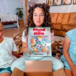 Three smiling girls sit on a living room floor, opening a box; the girl in the middle holds a book titled Blood Guts.