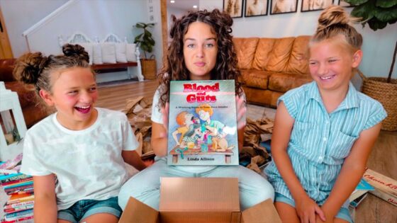 Three smiling girls sit on a living room floor, opening a box; the girl in the middle holds a book titled Blood Guts.