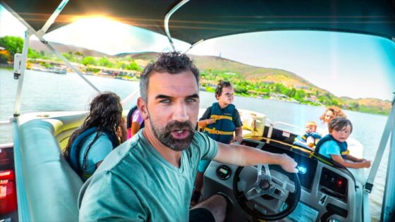 Man driving a motorboat while a group of children wearing life jackets sit in the back on a sunny lake. The man is reaching toward the steering wheel with a focused expression.