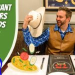 Man in a brown vest and blue plaid shirt lifts his hat and smiles at a cartoon girl with a backpack at a restaurant table; left side shows a green banner reading 'Restaurant Manners for Kids' with 'Cowboy Jack' logo; plate of food and a menu are on the table.