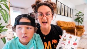 A woman with pink glasses and a child wearing a backward cap sit close together in a bright living room, looking surprised while holding a chart of colorful icons.