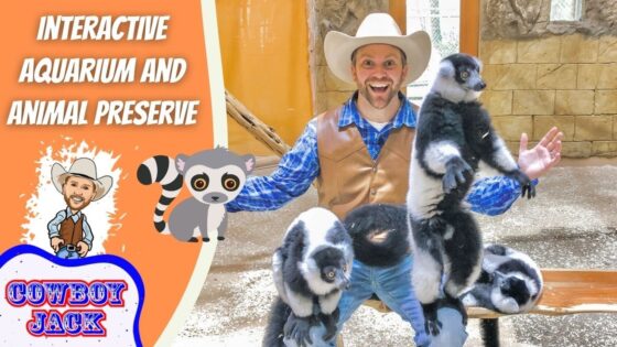 Smiling man in a cowboy hat with two lemurs perched on his arms, beside an orange sign that says 'INTERACTIVE AQUARIUM AND ANIMAL PRESERVE' and a 'COWBOY JACK' logo.