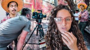 Behind-the-scenes shot of an outdoor film set: a woman with curly hair and pink glasses speaks to the camera while crew and equipment surround her.