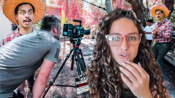 Behind-the-scenes shot of an outdoor film set: a woman with curly hair and pink glasses speaks to the camera while crew and equipment surround her.