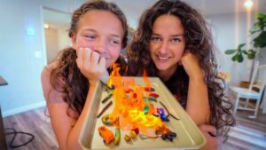 Two smiling young women lean close to a baking tray filled with colorful clay figures that are burning, watching the flames with curiosity.