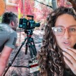 Behind-the-scenes of a video shoot: a woman with curly hair and pink glasses speaks to the camera, while a camera on a tripod and crew members work nearby.