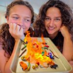 Two smiling girls watch a tray of burning colorful craft items during a science-like experiment in a bright room.
