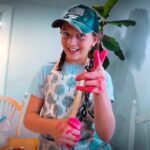 Girl with braids and a cap smiles at the camera while painting, her hands covered in bright pink paint, in a cozy dining room workspace.