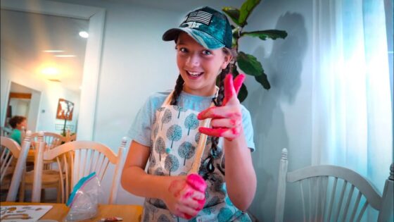 Girl with braids and a cap smiles at the camera while painting, her hands covered in bright pink paint, in a cozy dining room workspace.