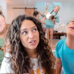 A woman with two young children close to the camera, all looking upward with smiles in a bright living room as other kids play in the background.