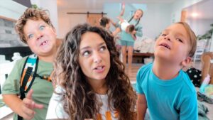 A woman with two young children close to the camera, all looking upward with smiles in a bright living room as other kids play in the background.
