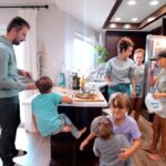 Family in a bright kitchen around an island, pizza on the board, adults and several children involved in cooking or serving food.