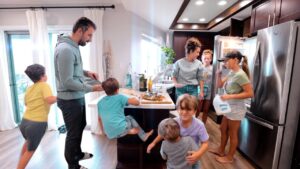 Family in a bright kitchen around an island, pizza on the board, adults and several children involved in cooking or serving food.