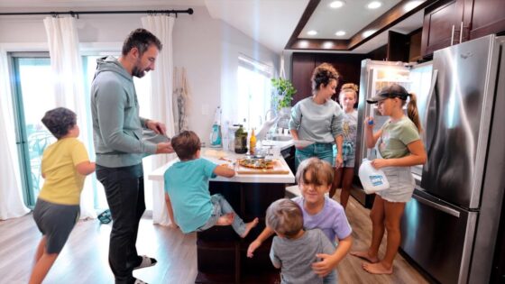 Family in a bright kitchen around an island, pizza on the board, adults and several children involved in cooking or serving food.