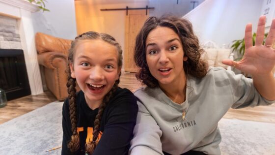 Two smiling women sit on a living room rug and wave at the camera, with a fireplace and couch in the background.