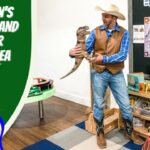 Man in cowboy hat and vest holds a toy dinosaur in a children’s museum play area, with dinosaur posters and books on shelves nearby.