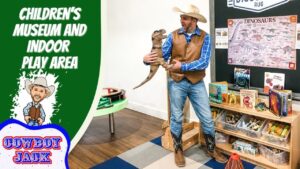 Man in cowboy hat and vest holds a toy dinosaur in a children’s museum play area, with dinosaur posters and books on shelves nearby.