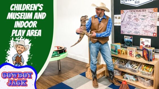 Man in cowboy hat and vest holds a toy dinosaur in a children’s museum play area, with dinosaur posters and books on shelves nearby.