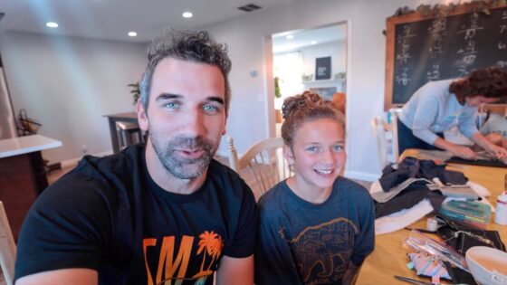 Man and young girl pose for a selfie at a craft table, smiling at the camera; sewing supplies and fabric are spread out, with a person sewing in the background in a bright room.