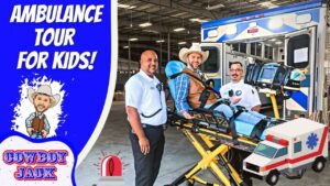 Paramedics pose with a patient on a stretcher beside an ambulance during a kids' ambulance tour event, with a blue panel reading 'Ambulance Tour for Kids' and cartoon cowboy.