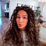 Woman with long curly hair takes a selfie in a bright home, while children play in the background.