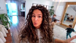Woman with long curly hair takes a selfie in a bright home, while children play in the background.