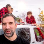 Family with plaid shirts decorating a white SUV with string lights in a park setting at sunset.