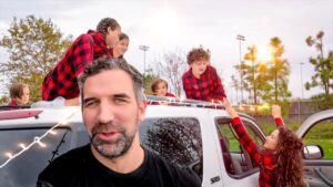 Family with plaid shirts decorating a white SUV with string lights in a park setting at sunset.