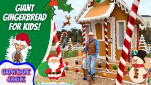 Father and child in cowboy outfits stand beside a giant gingerbread house at a holiday display with large candy canes and festive decorations.