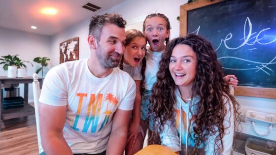 A smiling man and woman sit at a table while two excited girls stand behind them in a cozy room with a chalkboard and plants in the background.