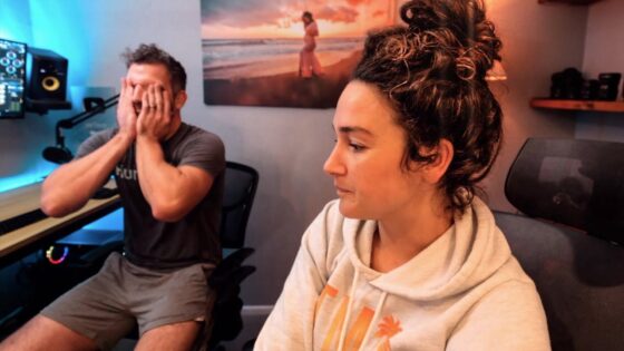 Man sits at a desk with hands covering his face while a woman in a hoodie sits nearby, in a home studio with blue and warm lighting.