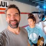 Two people inside a U-Haul truck take a selfie amid stacked boxes and packing materials in a cluttered moving setup.