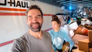 Two people inside a U-Haul truck take a selfie amid stacked boxes and packing materials in a cluttered moving setup.