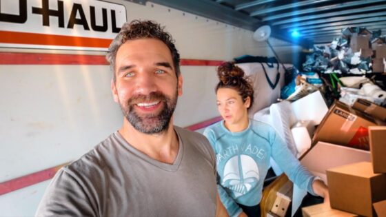 Two people inside a U-Haul truck take a selfie amid stacked boxes and packing materials in a cluttered moving setup.