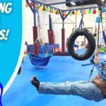 Cowboy-clad man swinging on a tire swing in an indoor play gym, with colorful equipment and a bold 'LEARNING OUR SENSES!' banner on the left dish side logo 'COWBOY JACK' visible.