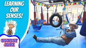 Cowboy-clad man swinging on a tire swing in an indoor play gym, with colorful equipment and a bold 'LEARNING OUR SENSES!' banner on the left dish side logo 'COWBOY JACK' visible.