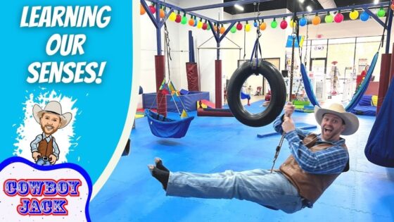 Cowboy-clad man swinging on a tire swing in an indoor play gym, with colorful equipment and a bold 'LEARNING OUR SENSES!' banner on the left dish side logo 'COWBOY JACK' visible.